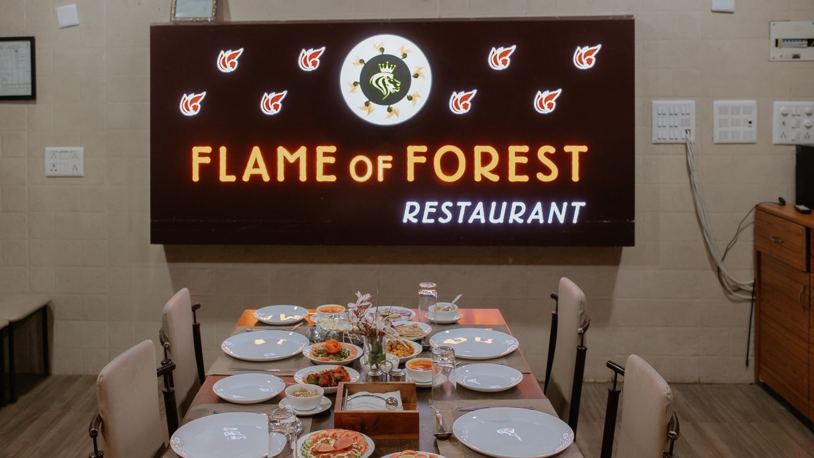 A clean, set dining table under the Flame of Forest Restaurant sign, awaiting diners, at Daksh Resort & Amusement Park, Sasan Gir.