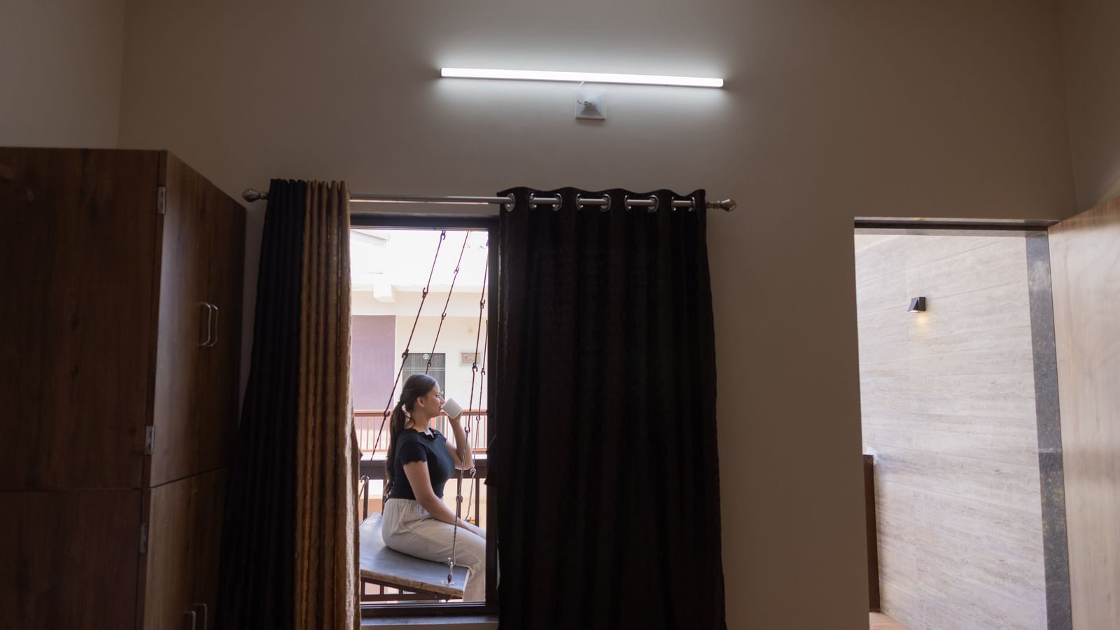 A shot of a lady sitting on a swing in a balcony taken from inside the room with the curtains and a wardrobe visible at Daksh Resort & Amusement Park, Sasan Gir.