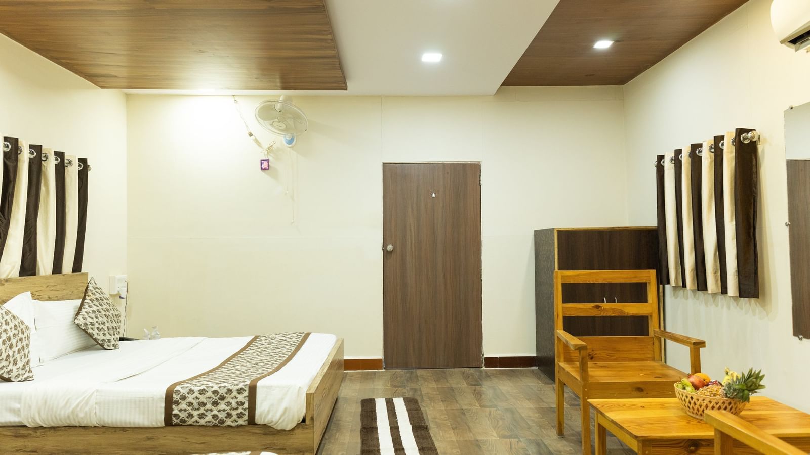 A bright room with a 2 beds, a wooden door, and 2 yellow wooden chairs with a table, at Daksh Resort & Amusement Park, Sasan Gir.