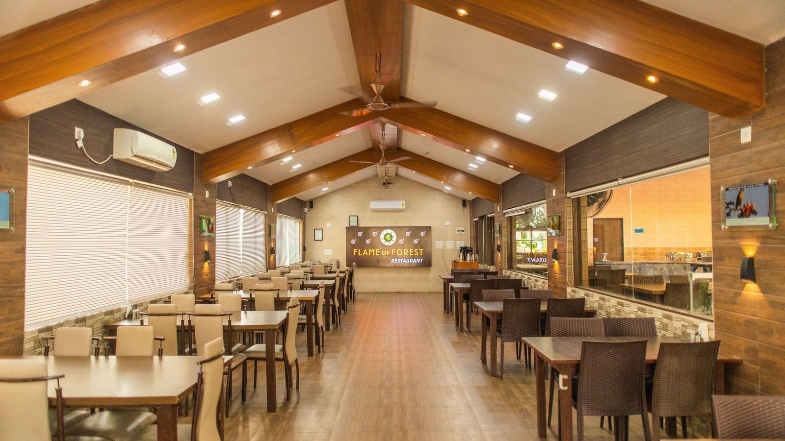 A spacious dining hall with wooden beams, ceiling lights, and rows of neatly arranged tables and chairs at Daksh Resort & Amusement Park's restaurant.
