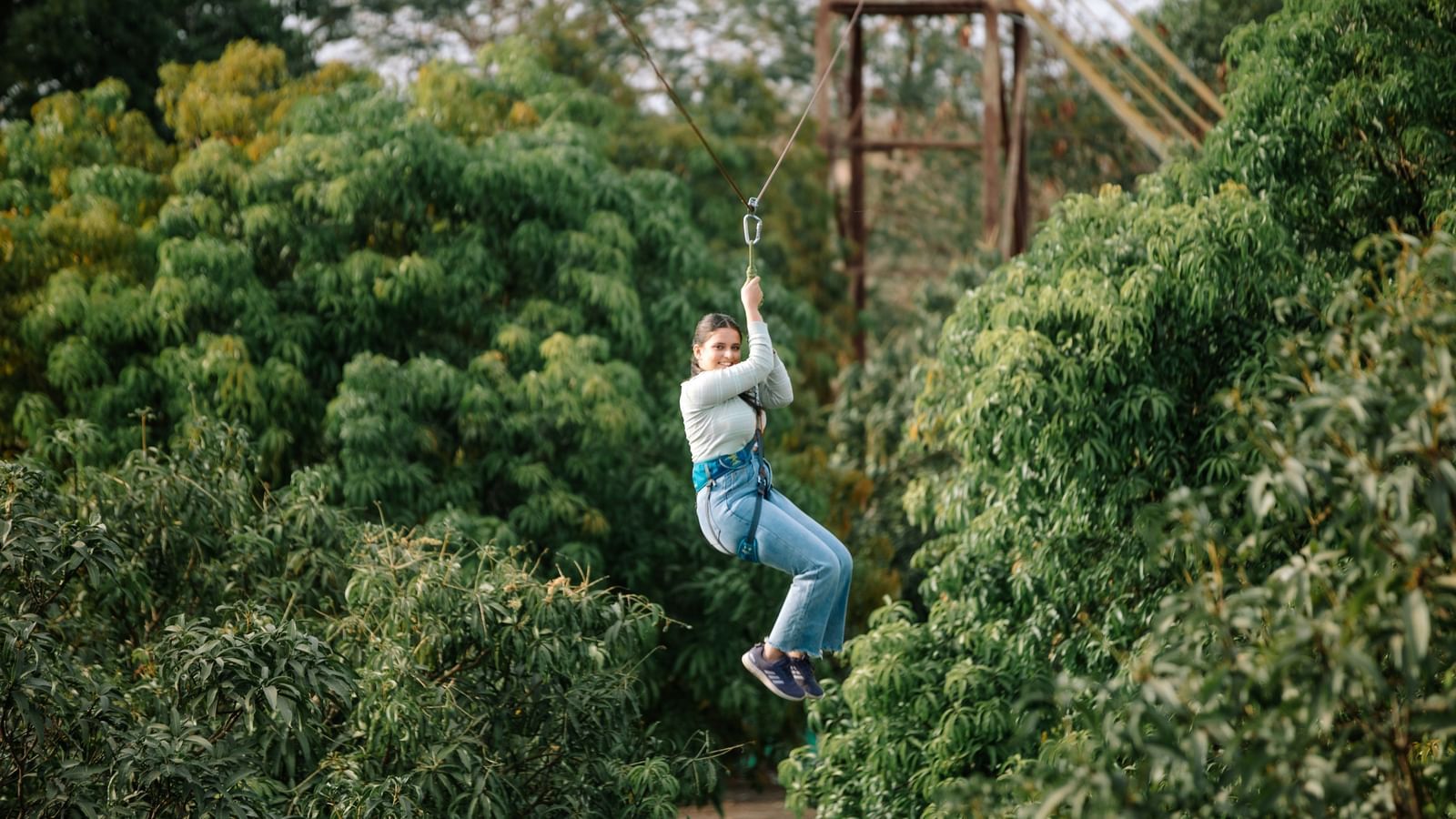 A person zip-lining through a dense green forest area, suspended mid-air by harness and cable at Daksh Resort & Amusement Park.