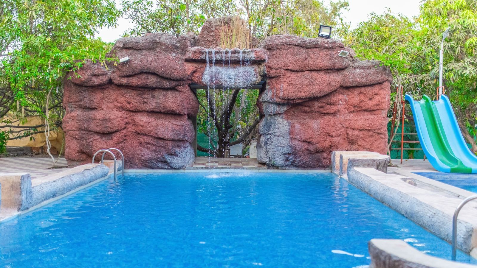 A swimming pool with a slide featuring a man-made waterfall at the background at Daksh Resort & Amusement Park.
