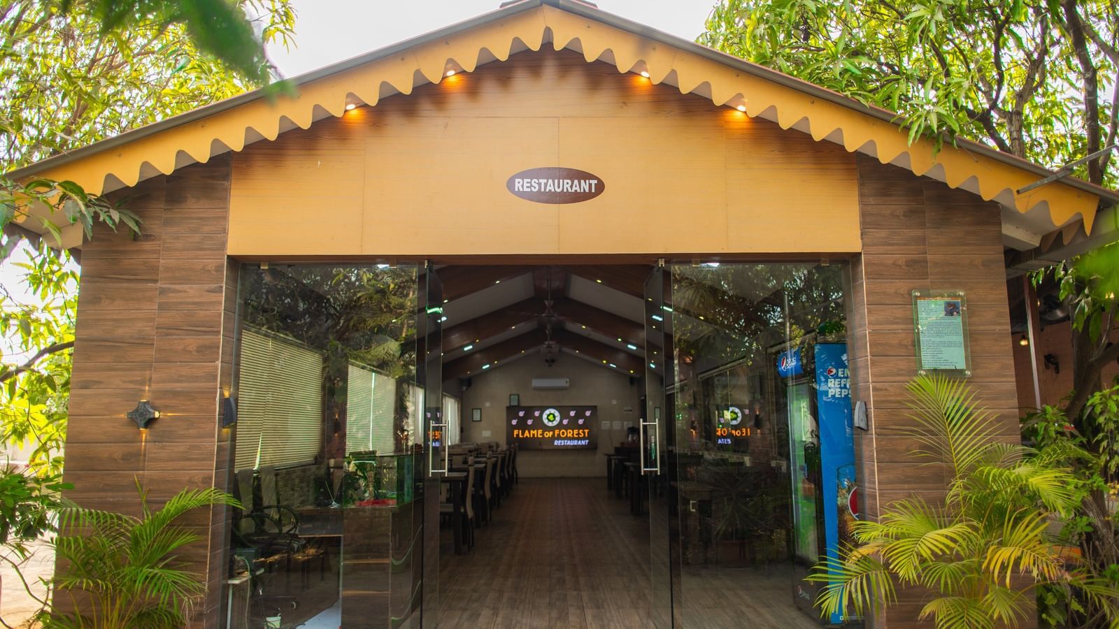 Cosy, glass-fronted restaurant featuring wooden interiors nestled in lush greenery at Daksh Resort and Amusement Park.