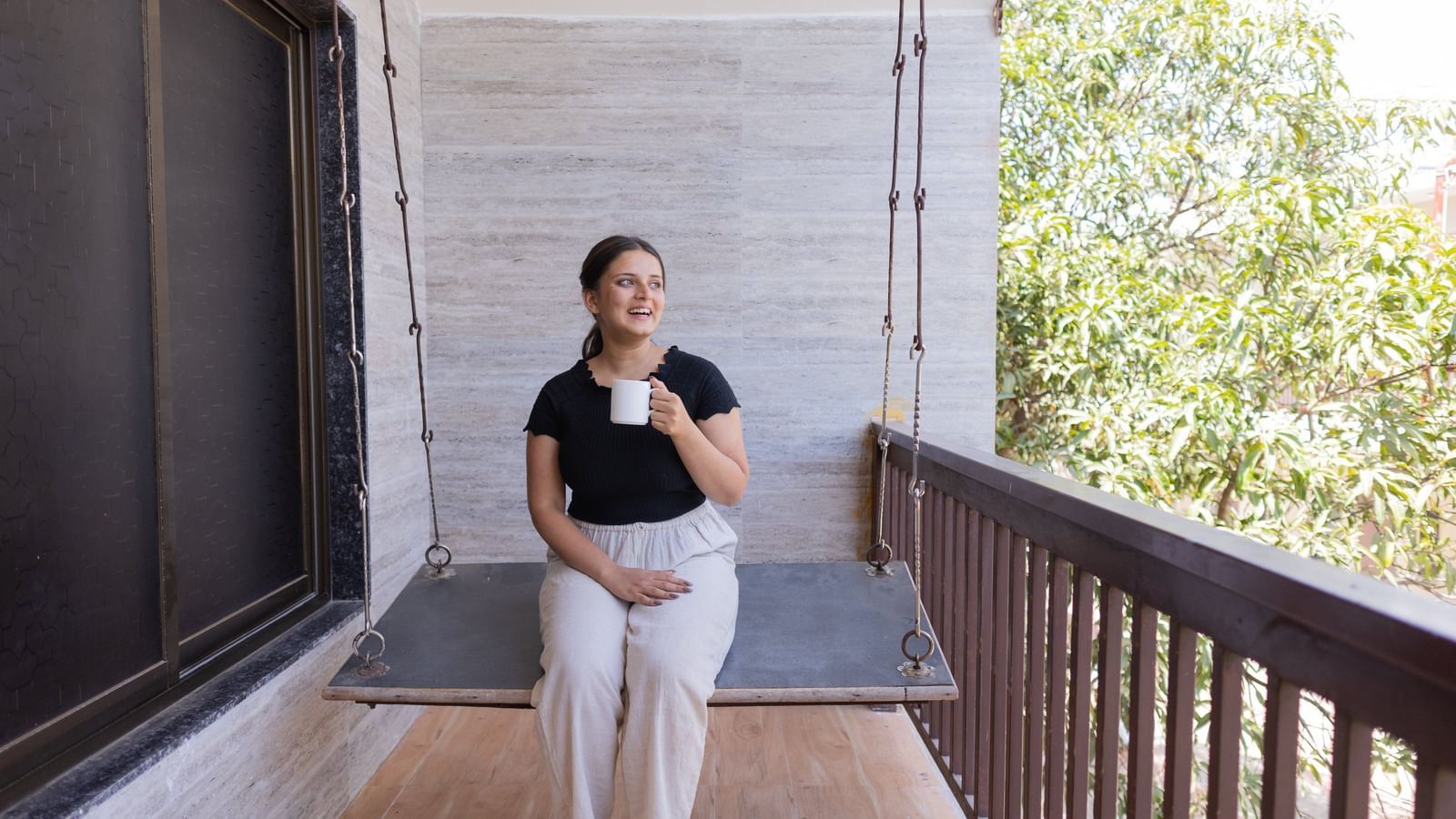 A woman sitting on a swing with a cup on her hand on a balcony with wooden railing beside a wall at Daksh Resort & Amusement Park.