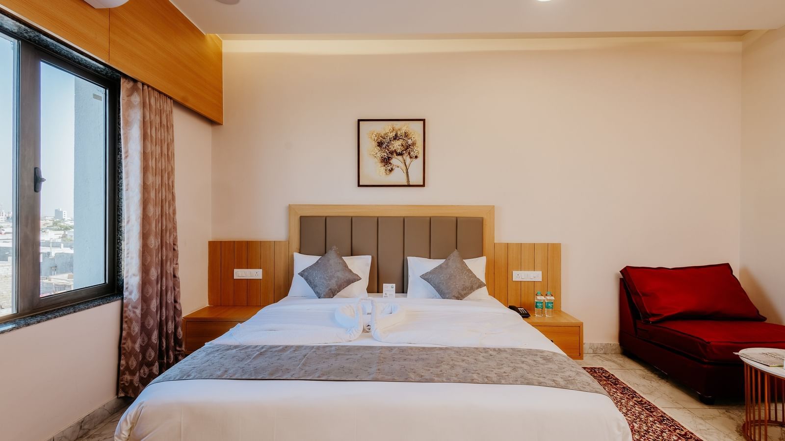 An inviting bedroom at Daksh The Grand Gowardhan, Dwarka, showcasing a spacious bed, bedside tables, and a vibrant red armchair in the corner.
