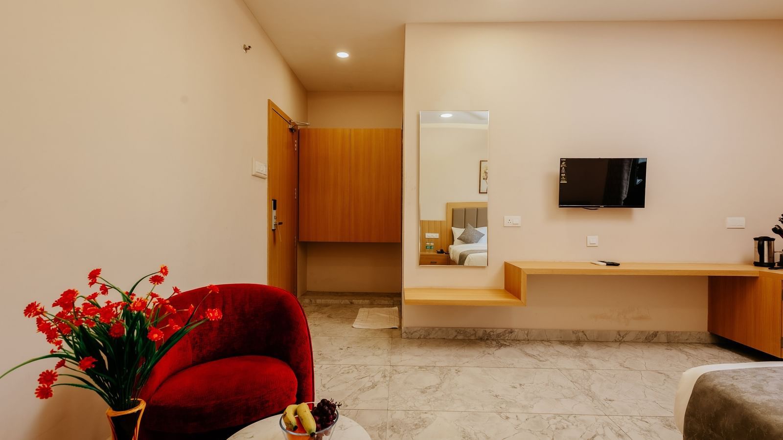 A spacious guest room interior at Daksh The Grand Gowardhan, Dwarka, with a comfortable bed, television, and a vibrant red armchair.
