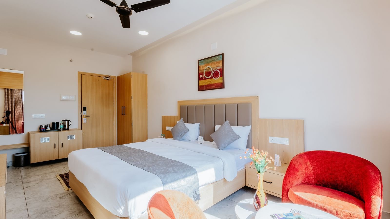 Corner view of a brightly lit bedroom at Daksh The Grand Gowardhan, Dwarka, showcasing a plush double bed, a vibrant red armchair along with a coffee table in front of it.