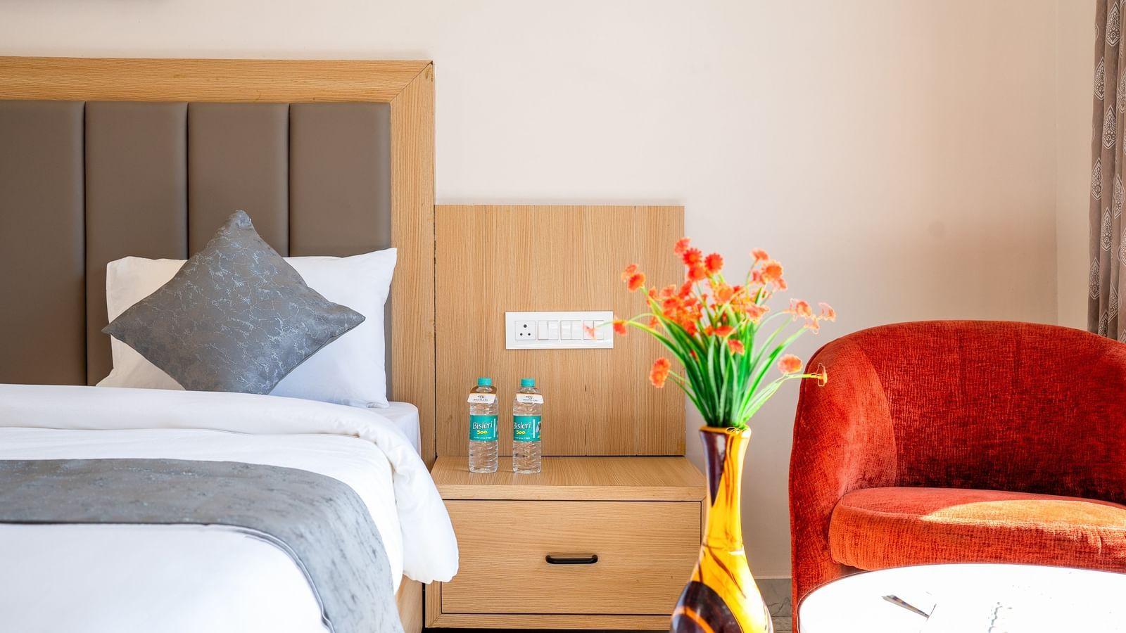 A closeup of a neatly made bed along with a bedside table and an elegant red armchair in a bedroom at Daksh The Grand Gowardhan, Dwarka.