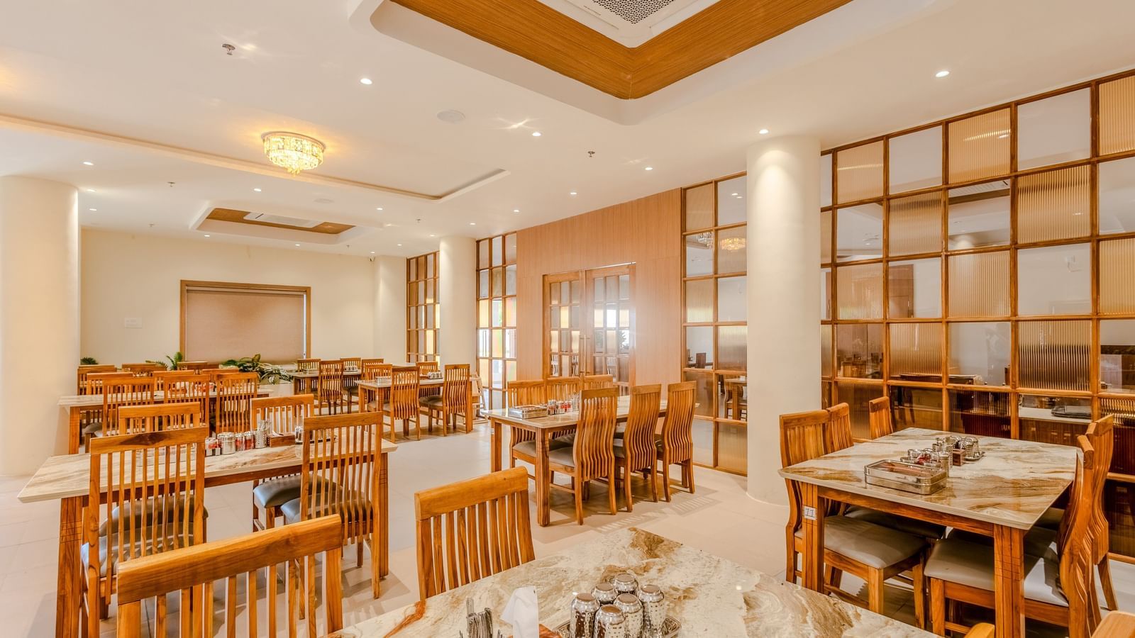 A wide view of the restaurant at Daksh The Grand Gowardhan, Dwarka, showing multiple dining tables and decorative shelving along with central air-conditioning.