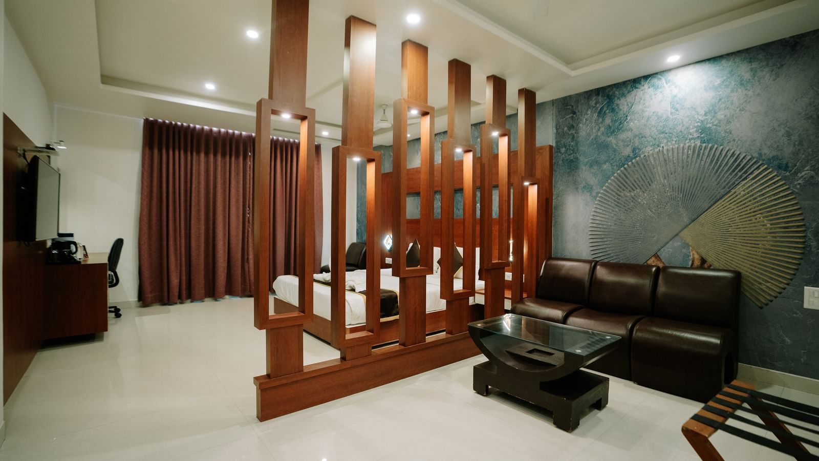 Wide view of a sitting area separated from the bedroom by a stylish, tall wooden slat partition. Dark leather sofa at Daksh The Madhuvan Suites, Dwarka.