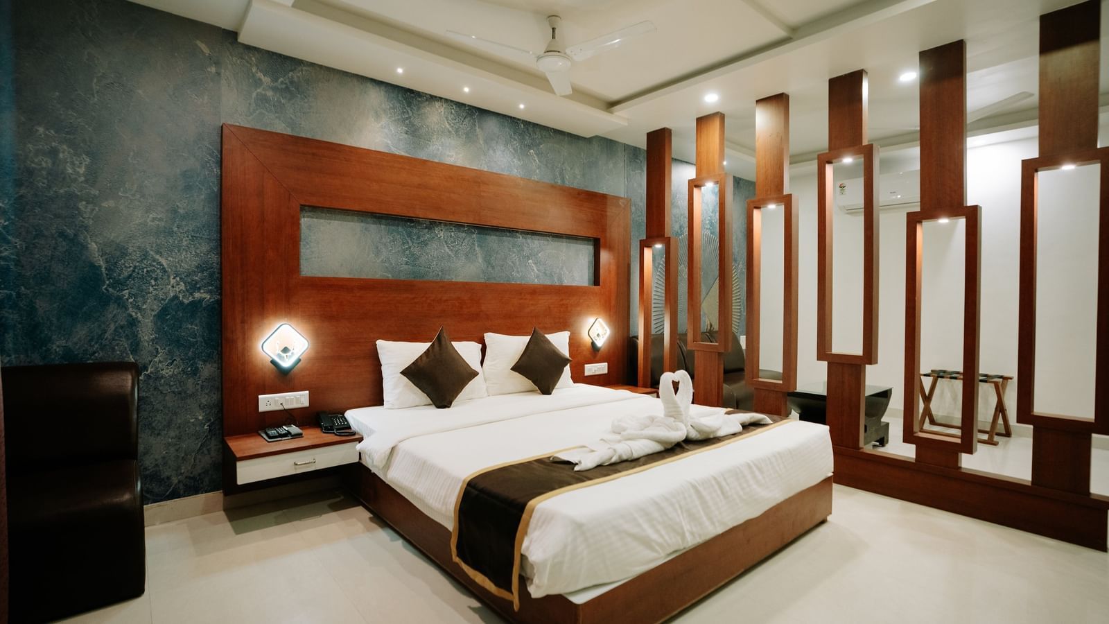 Double bed with dark wood frame and headboard, set against a blue textured feature wall, with decorative wooden partitions at Daksh The Madhuvan Suites, Dwarka.
