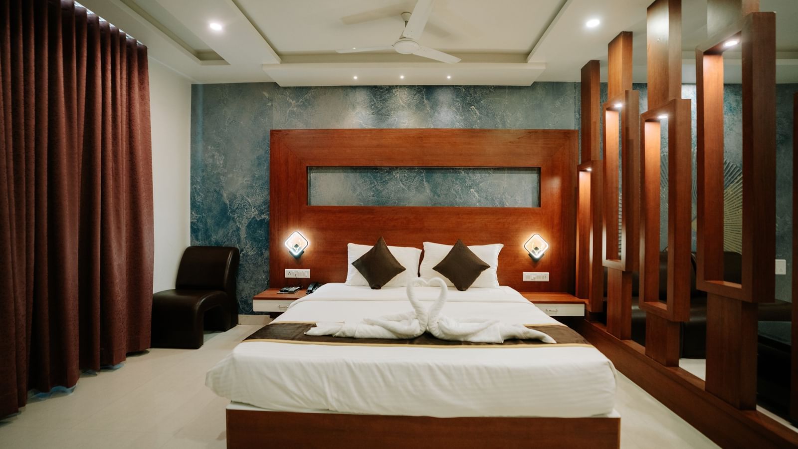 Close-up of the double bed flanked by wooden slats and warm bedside lighting, highlighting the dark wooden headboard at Daksh The Madhuvan Suites, Dwarka.