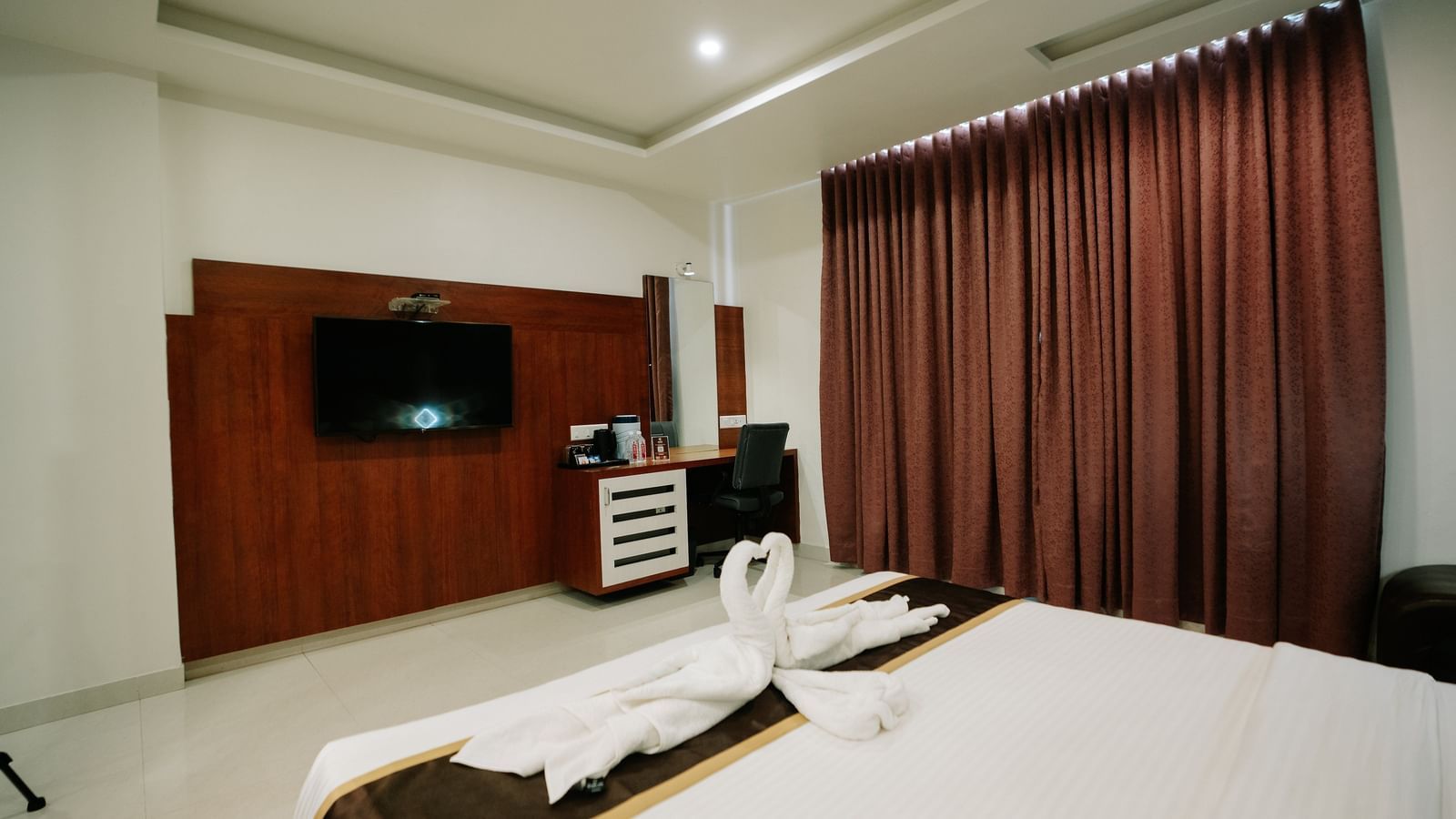 Well-appointed hotel room with a large bed, brown bedding, towel swans, rich burgundy curtains, and a TV unit at Daksh The Madhuvan Suites, Dwarka.