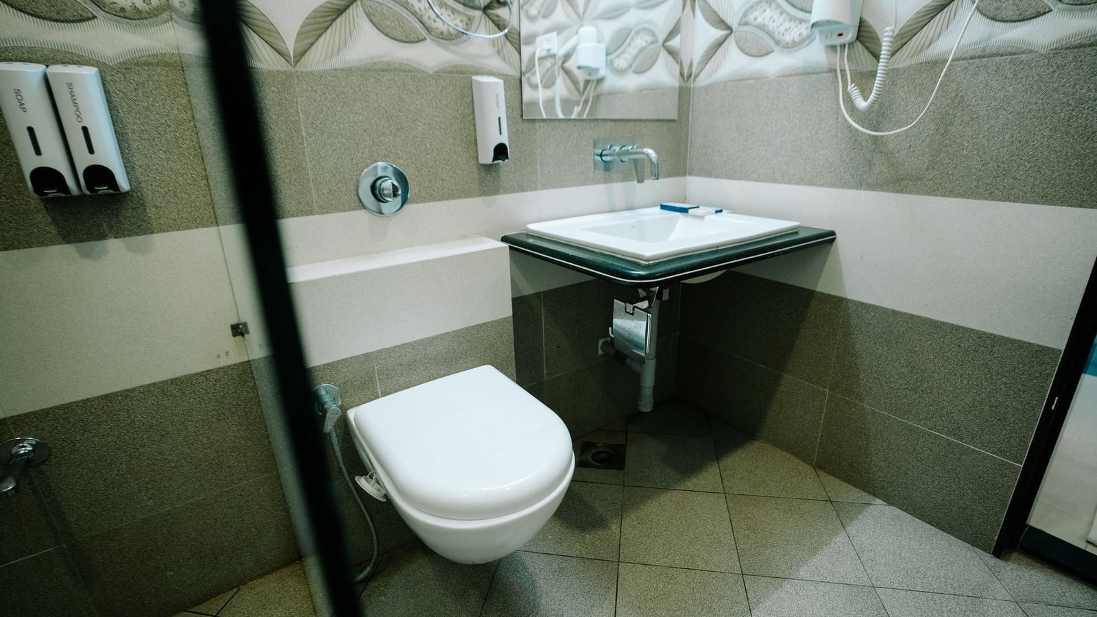 View of the clean, modern bathroom showing the square wall-mounted basin and the toilet, framed by light-coloured patterned tiles at Daksh The Madhuvan Suites, Dwarka.