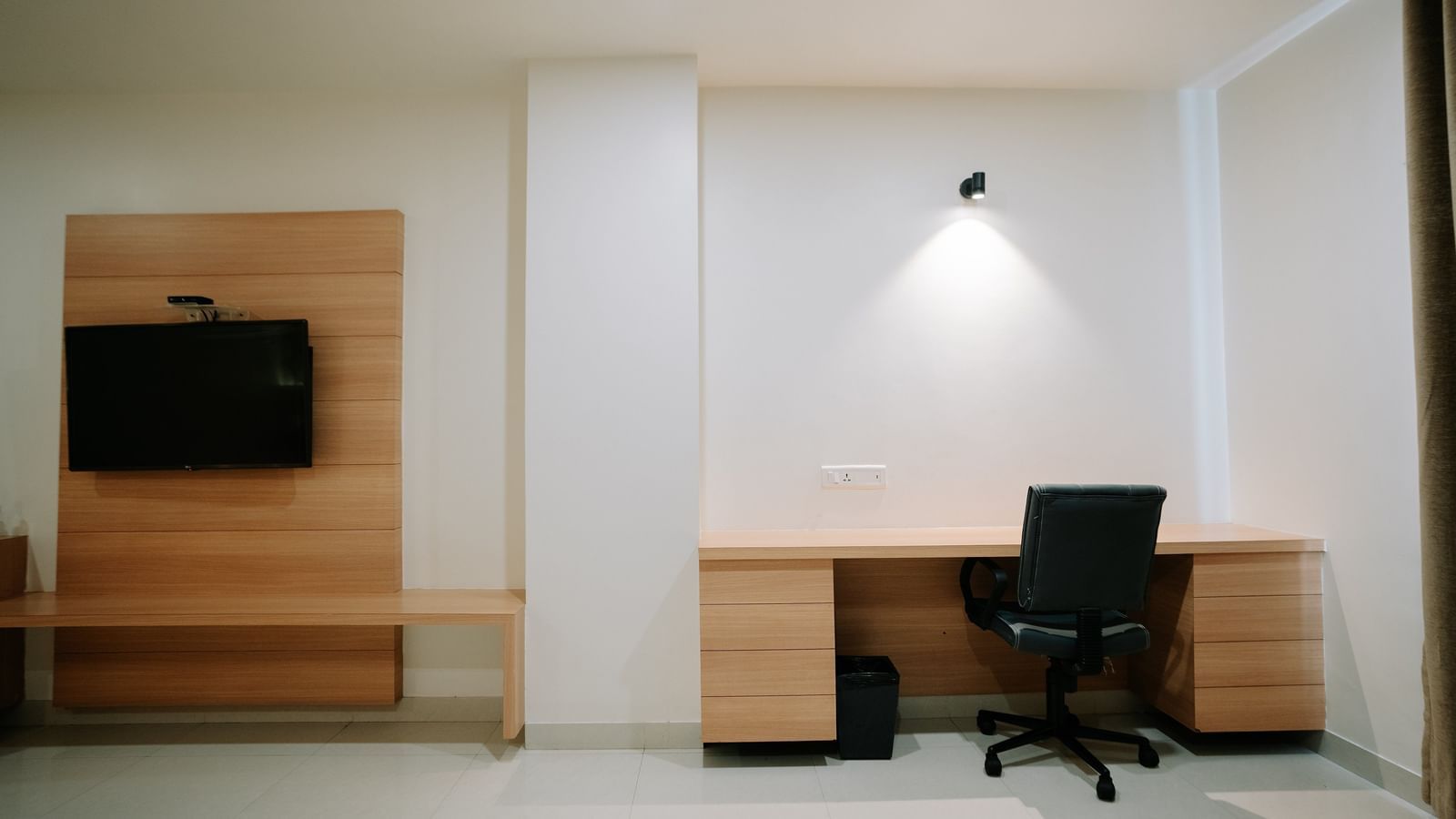 Minimalist wooden desk area with a wall-mounted TV, white paneling, spot lighting, and a black office chair at Daksh The Madhuvan Suites, Dwarka.