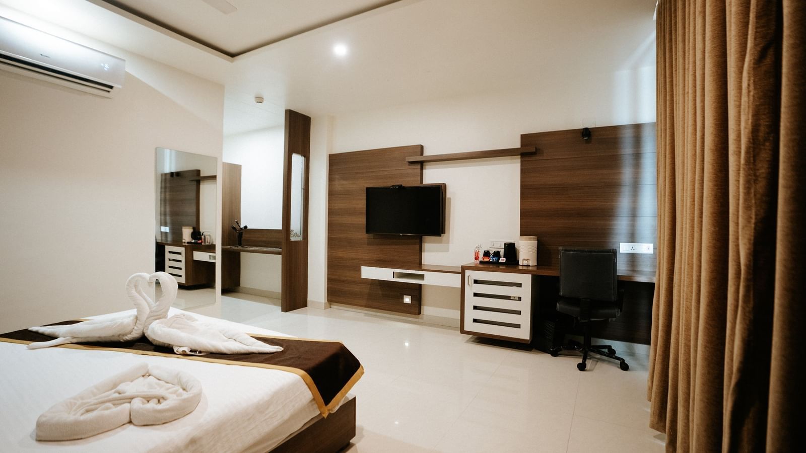 Panoramic view of the upscale hotel room, showing the bed, dark wood furniture, media console, and rich brown curtains at Daksh The Madhuvan Suites, Dwarka.