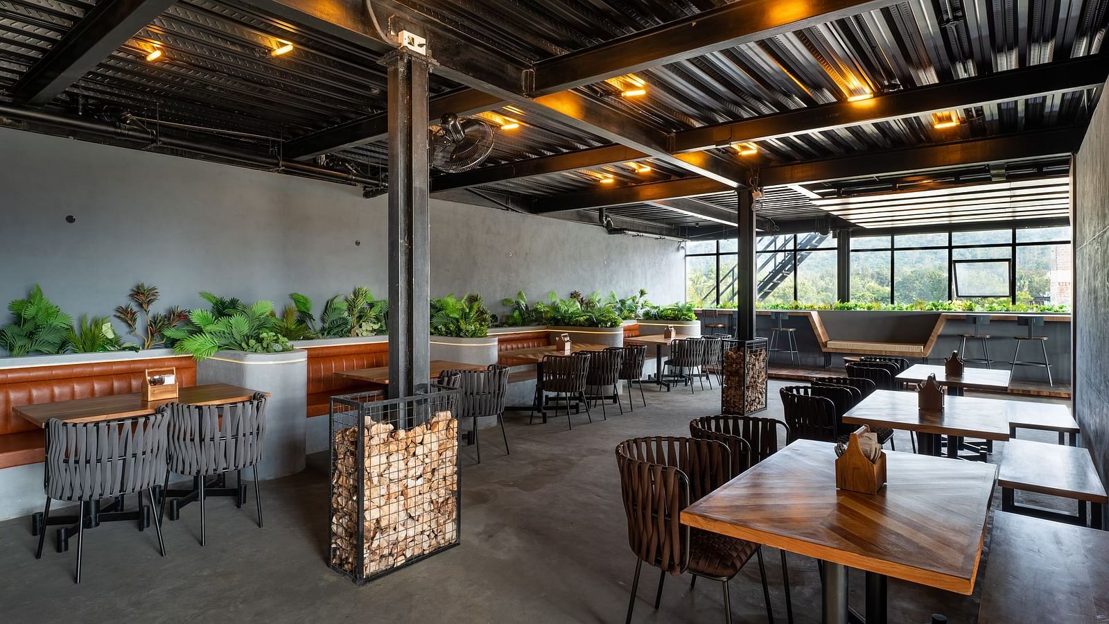 A modern open-air restaurant with wooden tables, cushioned seating, industrial-style ceiling, and lush greenery, offering a stylish dining experience at TGI Livancia Hillside, JC Nagar, Mysore.