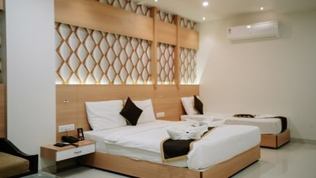 Close-up view of the bed and bedside table. White linen, dark pillows, and a wooden headboard with geometric lattice detail at Daksh The Madhuvan Suites, Dwarka.