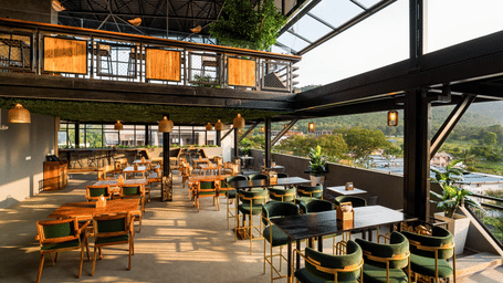 A spacious rooftop restaurant with elegant wooden furnishings, green velvet chairs, and panoramic views of the hills, bathed in warm natural light at TGI Livancia Hillside, JC Nagar, Mysore.