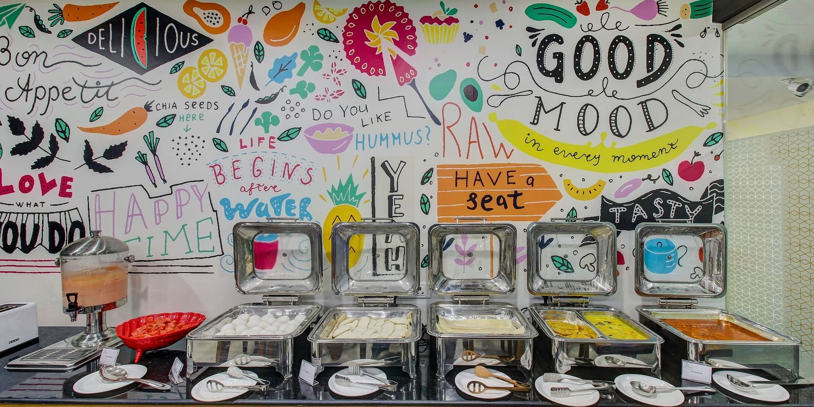 A hotel buffet corner with a wall mural saying Good Mood Bon Appétit and various other cheerful phrases alongside a spread of breakfast items ready for guests.