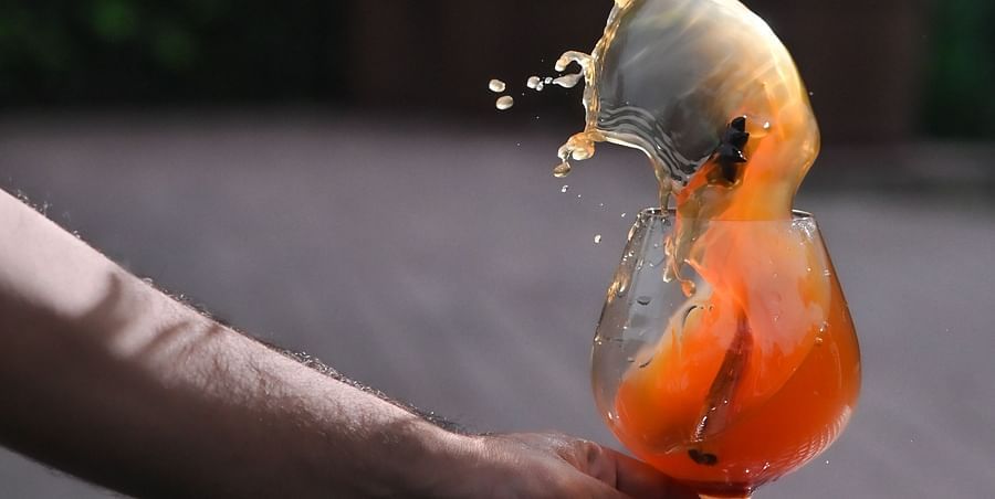 Action shot of a hand holding a glass of orange-coloured drink with ice, as the liquid splashes dramatically out of the glass, set against an outdoor background with greenery - Ananta Spa and Resort, Pushkar.