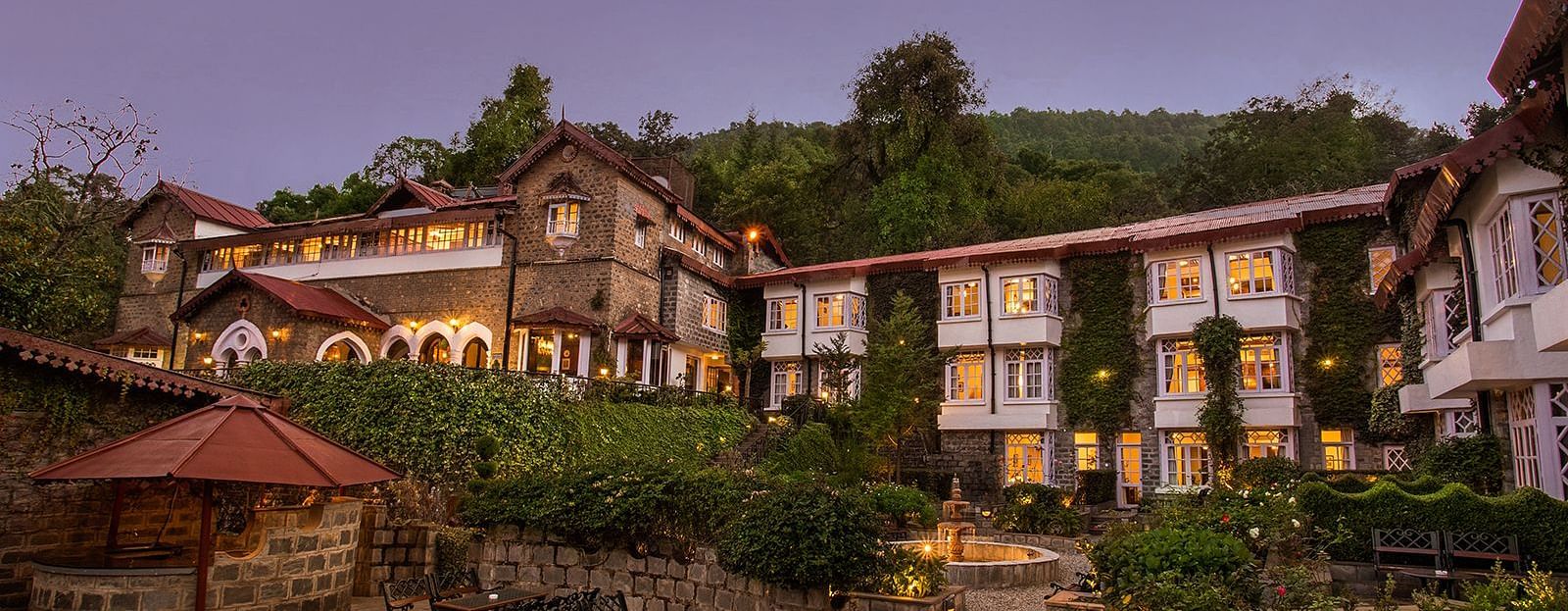 A scenic view of a the naini retreat, nainital, The resort features multiple buildings with stone exteriors and white trim. The surrounding landscape includes mountains, trees, and a winding pathway.