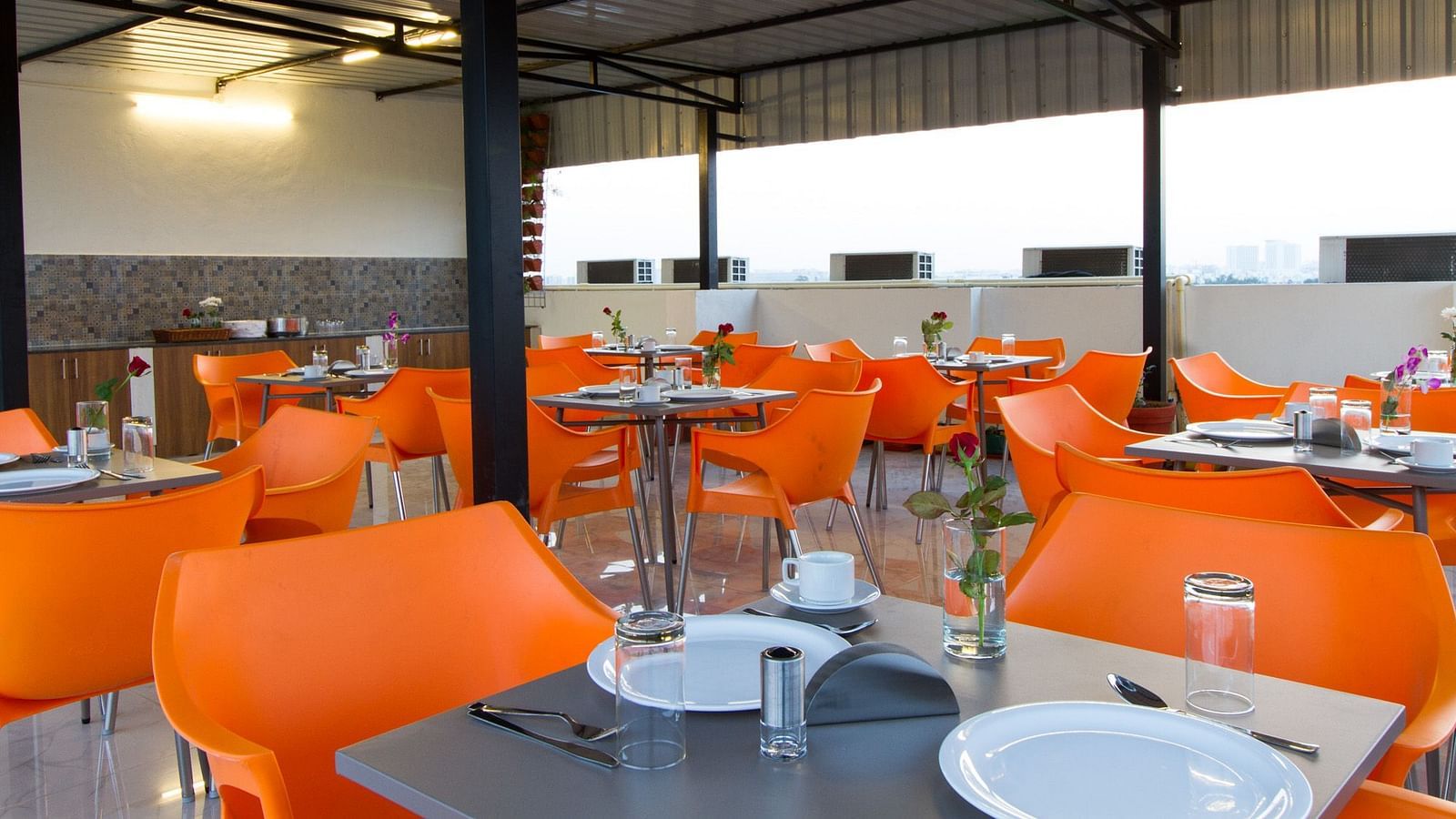 An outdoor dining space under a canopy offers numerous tables with bright orange seating at Sanctum Suites Whitefield, Bangalore.
