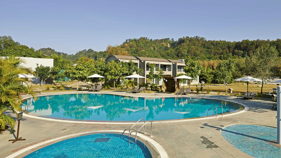 A large, inviting swimming pool with blue water sits in a resort area, surrounded by sun loungers, umbrellas, and buildings with green hills behind.
