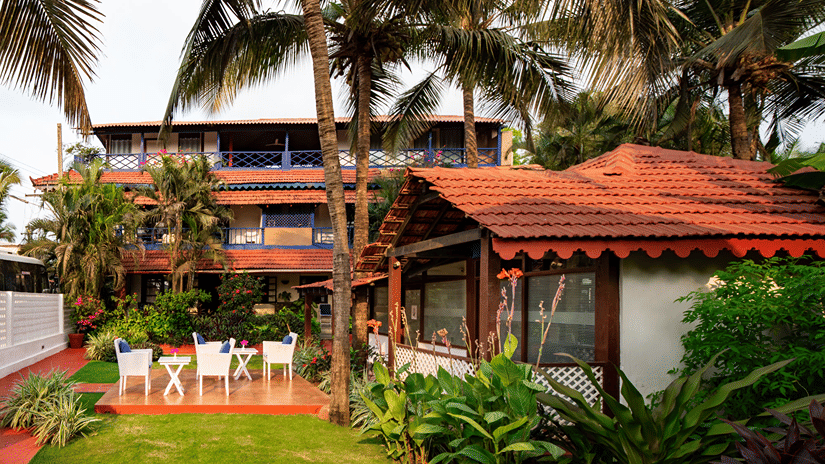 Red-roofed outdoor cottage surrounded by palm trees, tropical plants and garden seating.