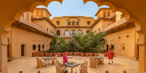 The central courtyard of Neemrana's Tijara Fort Palace, viewed through an ornate archway, showcases seating areas and lush greenery within the historic structure.