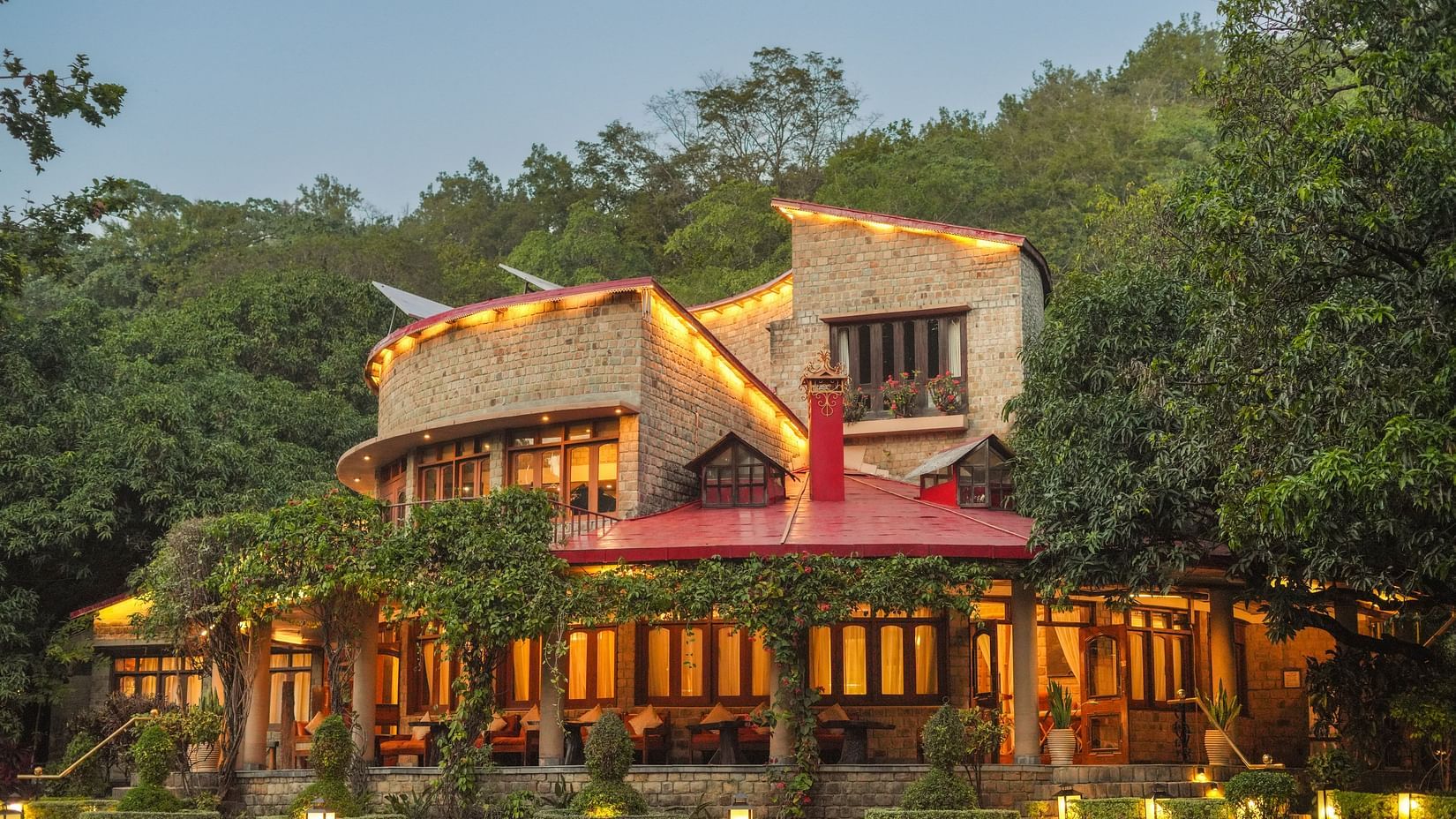 A multi-level building with lights, nestled in a forested hillside at dusk.