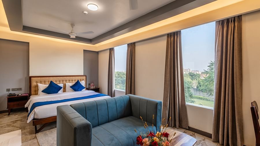 Deluxe King Room at Clarks Inn Suites Jaipur featuring a plush bed by the window under warm lighting with a sofa at its foot along and a coffee table