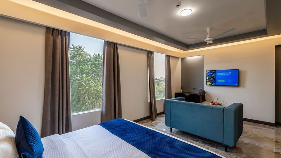 Deluxe King Room at Clarks Inn Suites Jaipur featuring a plush bed facing the TV mounted on the wall in front of the sofa at its foot and large windows with a lush view