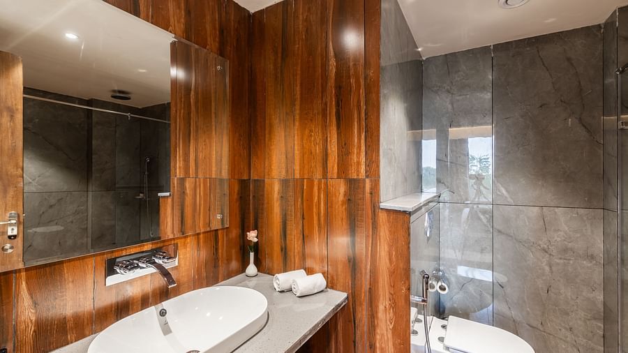 Bathroom of Deluxe King Room at Clarks Inn Suites Jaipur with a wooden finish, featuring a sleek countertop basin, a large mirror, a glass-enclosed shower area, and ambient lighting for a luxurious feel