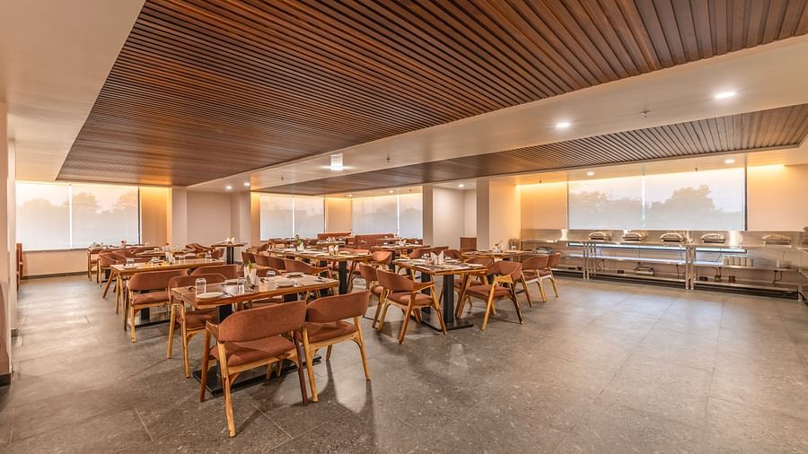 A restaurant with a buffet counter, wooden ceiling panels, neatly arranged tables, brown chairs and large windows with blinds at The Bridge - Clarks Inn Suite Jaipur