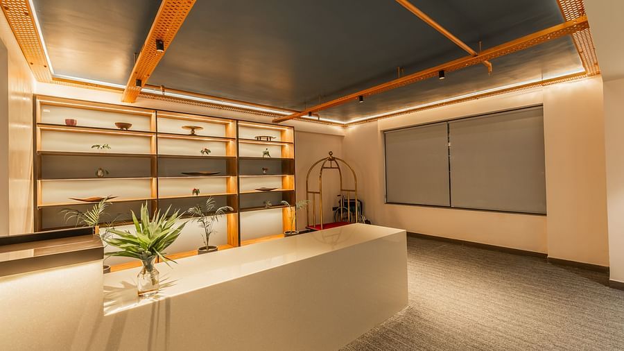 A well-lit reception at Clarks Inn Suites Jaipur, Ajmer Road featuring a reception desk with a monitor and a plant on it and showpieces behind the desk on the shelves