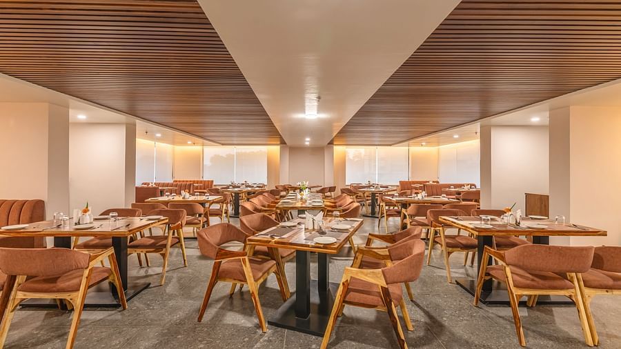 A spacious restaurant of The Bridge with wooden ceiling panels, modern lighting, and neatly arranged tables with brown chairs and elegant table settings at Clarks Inn Suites Jaipur