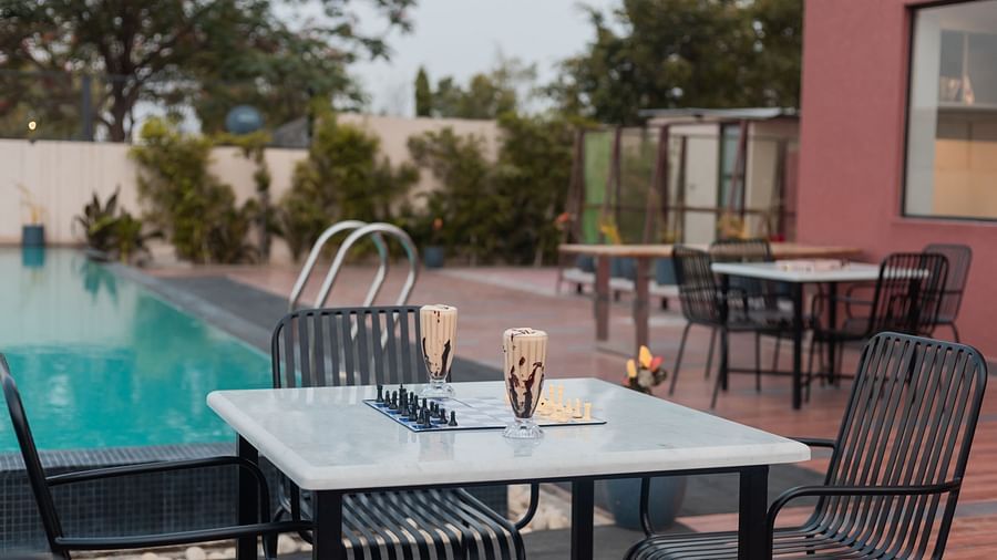 An expansive outdoor patio at Clarks Inn Suites Jaipur, Ajmer Road with numerous dining tables and chairs under a long, slatted pergola, surrounded by lush greenery and a bright sky.