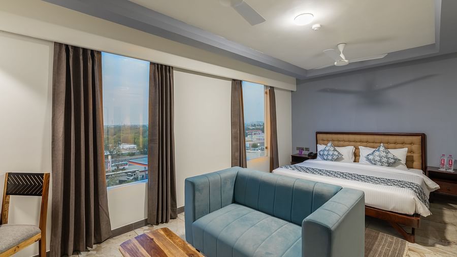 Clarks Inn Suites Jaipur, Ajmer Road featuring room with large windows, king size bed, blue sofa, wooden furniture, ceiling fans, and city view outside the curtains.