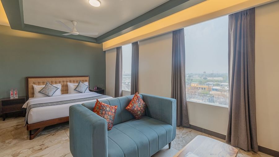 Deluxe King Room at Clarks Inn Suites Jaipur featuring a cosy bed, modern blue sofa, large windows, elegant decor, warm lighting, and a scenic view