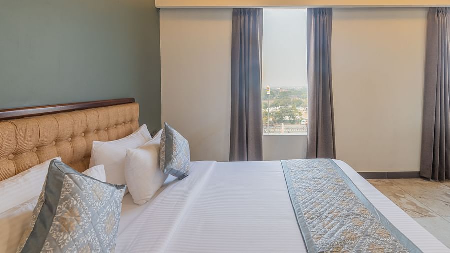 Deluxe King Room at Clarks Inn Suites Jaipur with a plush king-size bed, decorative pillows, soft linens, large window with curtains, and warm ambient lighting