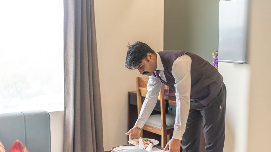 Room service staff member in the Deluxe King Room, Clarks Inn Suites Jaipur in a smart uniform is setting up a tray of food in a well-lit, stylish room with elegant furnishings and warm decor
