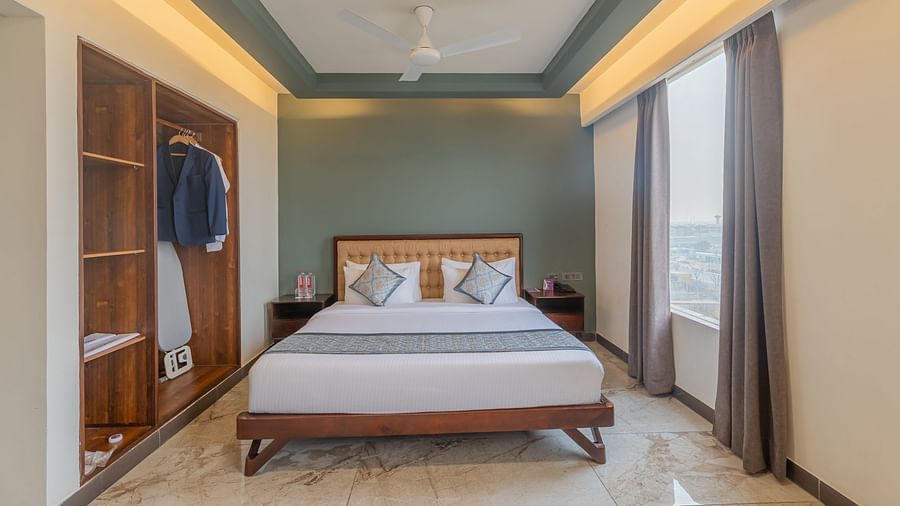 Deluxe King Room at Clarks Inn Suites Jaipur featuring a king size bed, open wardrobe with hanging clothes, large windows with curtains, soft ambient lighting, marble flooring, and a modern, elegant design