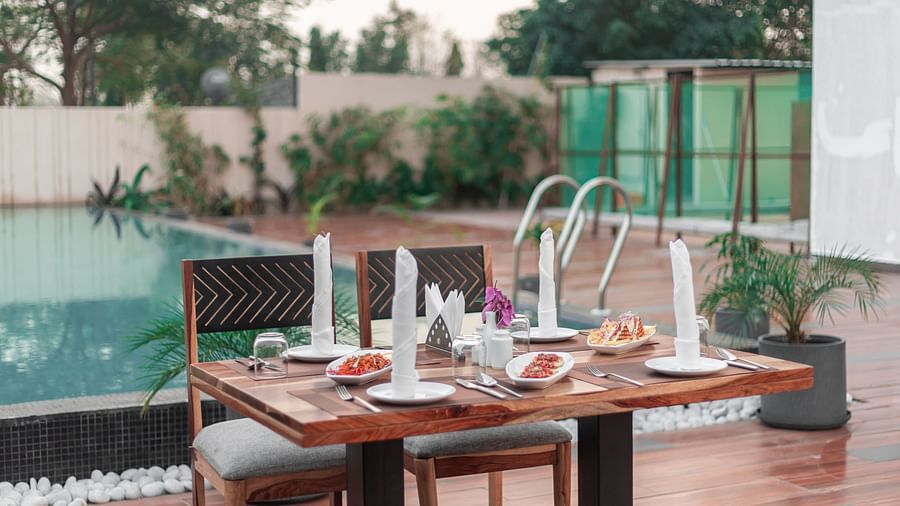 Poolside dining setup with a wooden table, stylish chairs, and plated dishes amid greenery at The Bridge, Clarks Inn Suites Jaipur