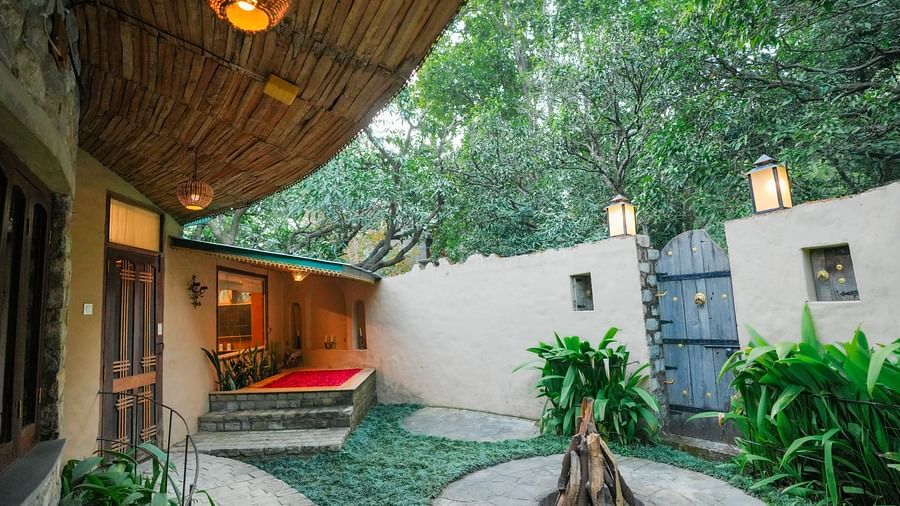 An outdoor courtyard with a stone pathway, plants, and a building with a thatched roof - The Den Corbett Resort & Spa, Kumeria.