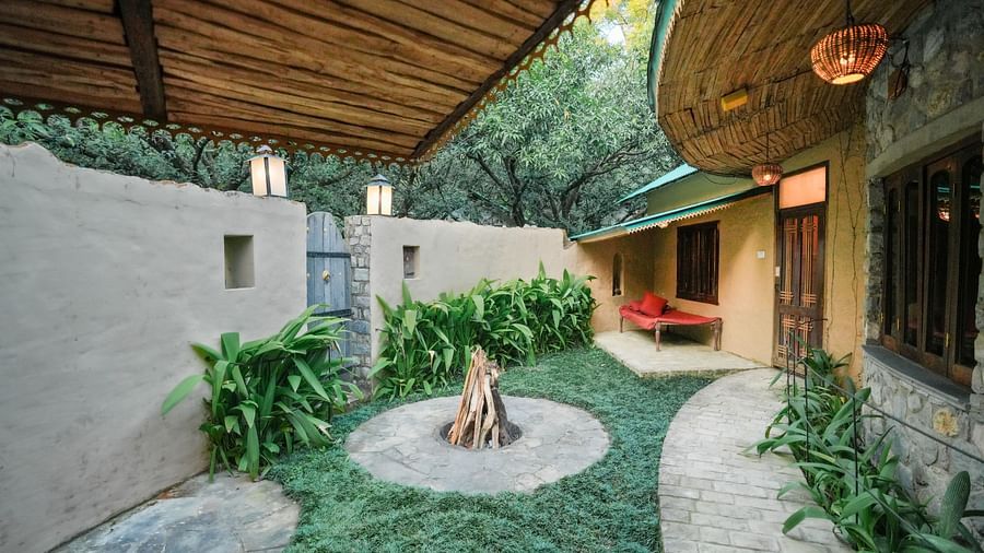 An outdoor courtyard with a stone pathway, plants, and a building with a thatched roof - The Den Corbett Resort & Spa, Kumeria.