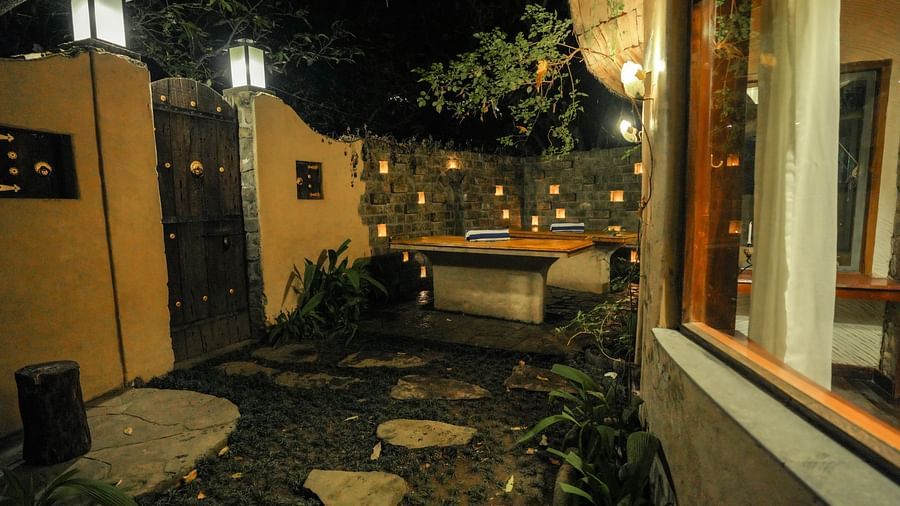 An outdoor patio with a stone pathway, plants, and a building with warm lighting at night - The Den Corbett Resort & Spa, Kumeria.