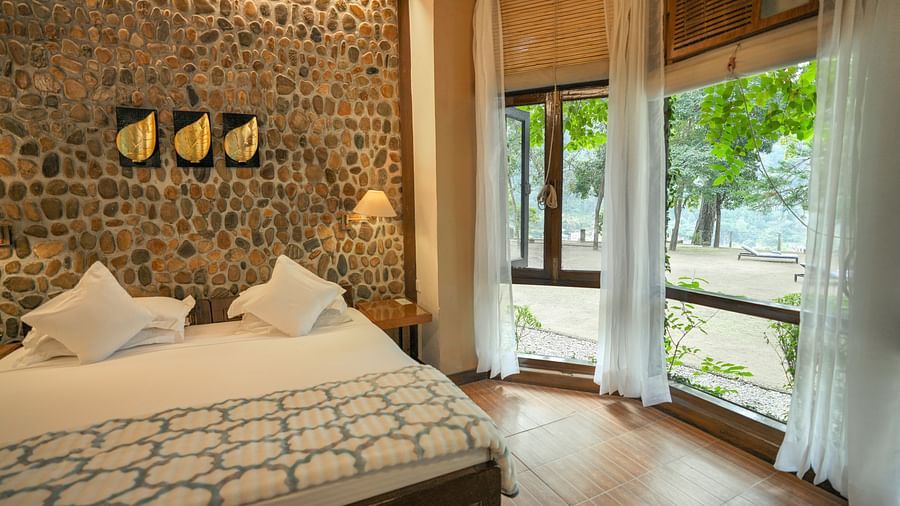 A bedroom with a bed, a stone wall, and large windows - The Den Corbett Resort & Spa, Kumeria.
