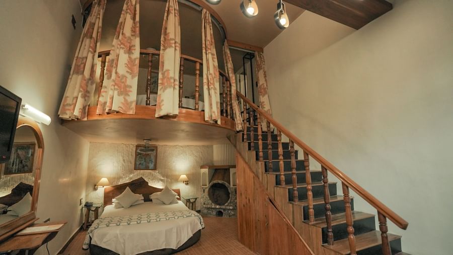 A bedroom with a bed, a rug, and warm lighting, interior setting with wooden elements and stairs - The Den Corbett Resort & Spa, Kumeria.