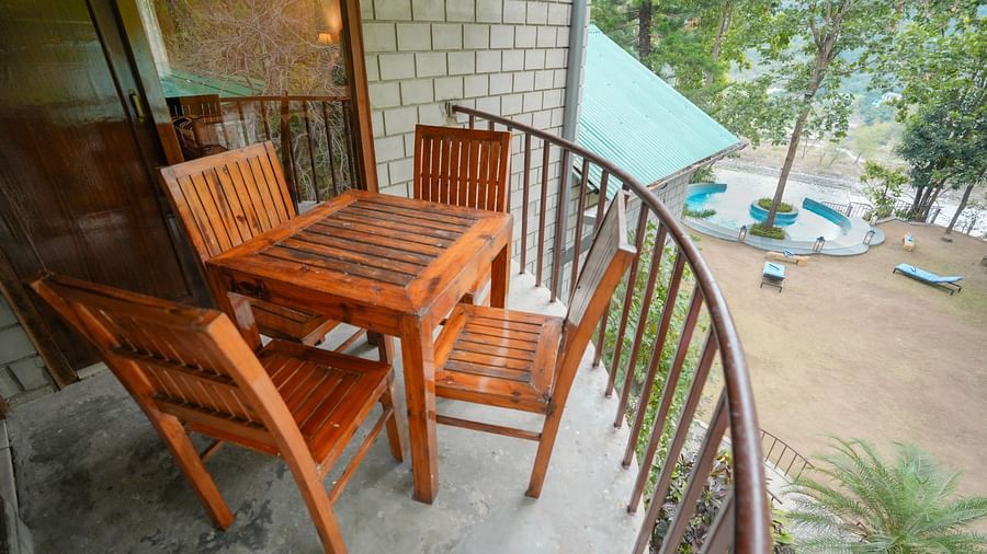 An outdoor balcony with a wooden table and chairs, overlooking a body of water - The Den Corbett Resort & Spa, Kumeria.